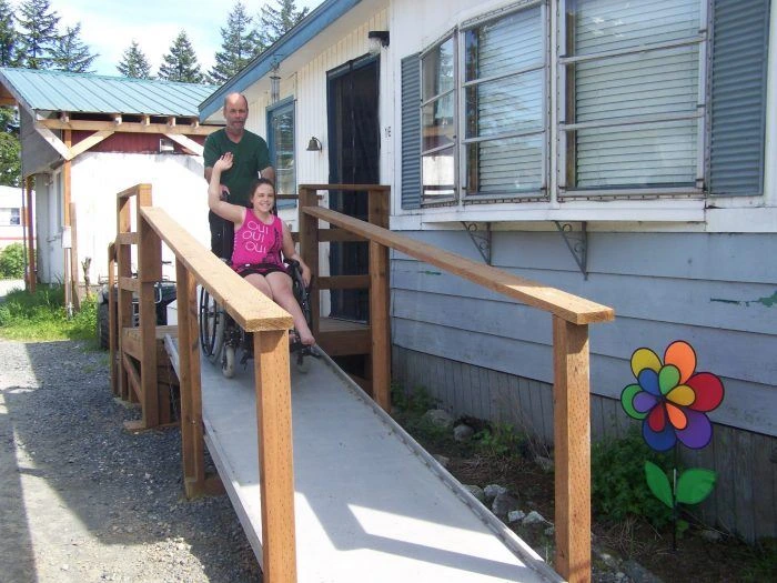 Woman in wheelchair smiling and waving as she rolls down an accessible ramp from a light blue house.